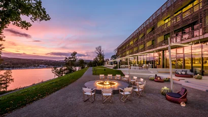 Abendliche Terrasse mit Feuerstelle und Stühlen neben einem modernen Gebäude am See