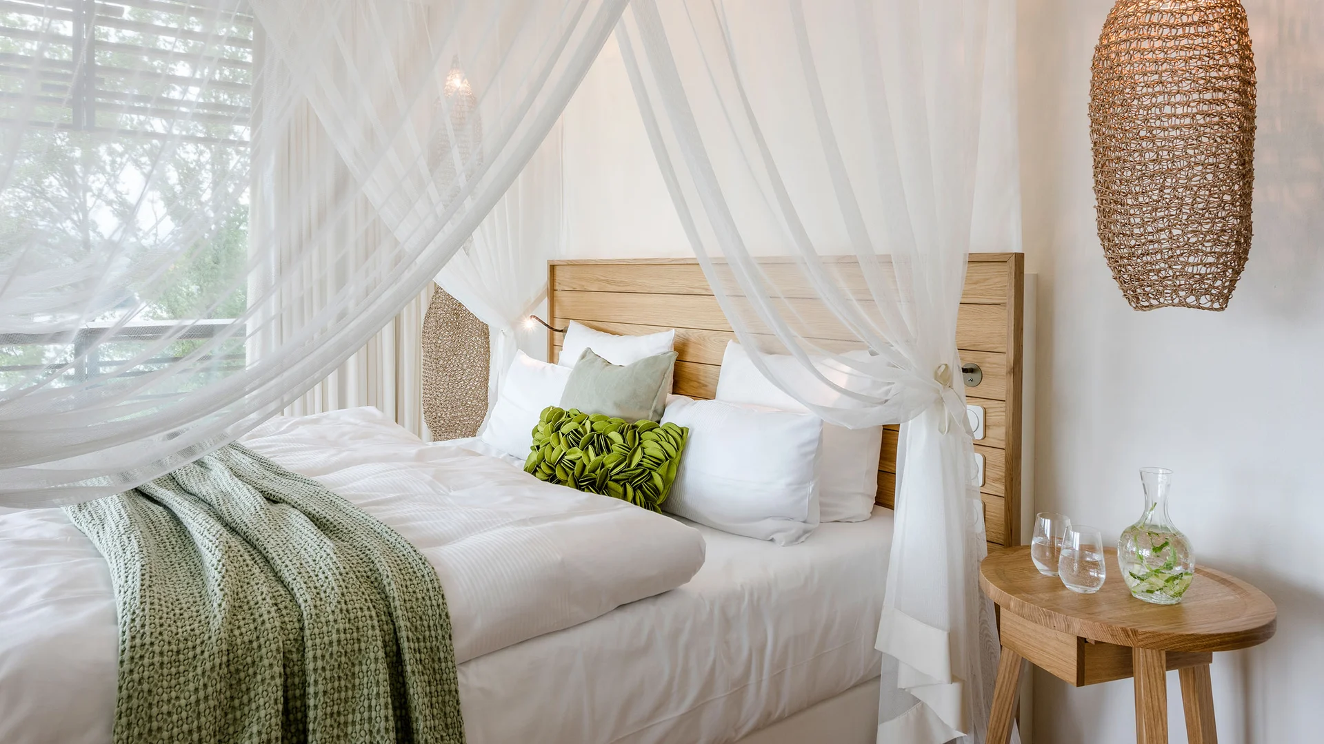 Helles Schlafzimmer mit Himmelbett, grünen Kissen und Holztisch mit Wasserkrug