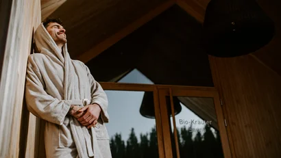 Mann in Bademantel lehnt an Sauna-Holzwand und lächelt entspannt