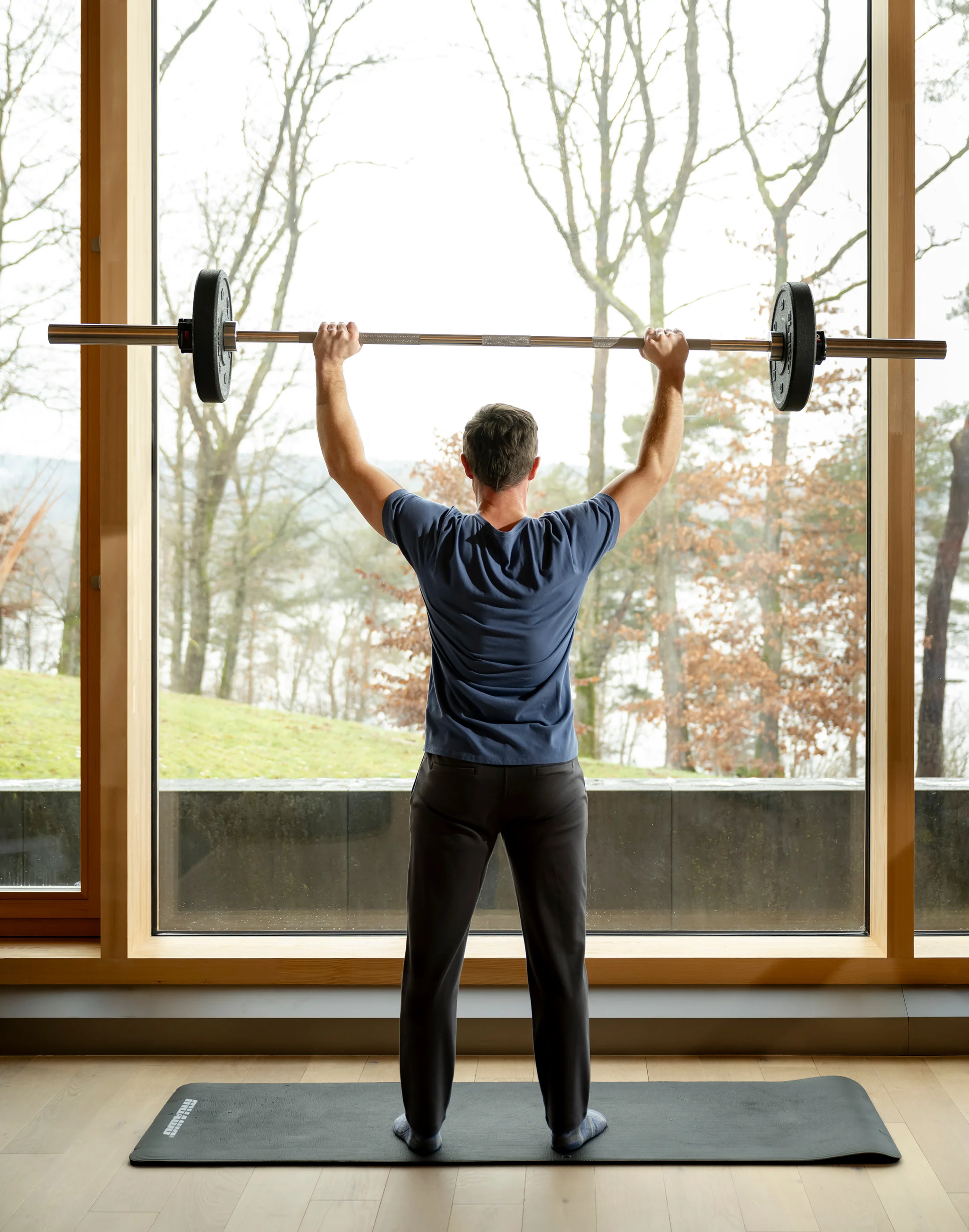 Mann hebt Langhantel vor großem Fenster mit Naturblick