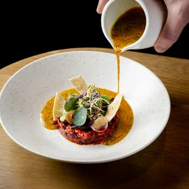 Hand gießt Sauce über angerichteten Salat in weißem Teller auf Holztisch