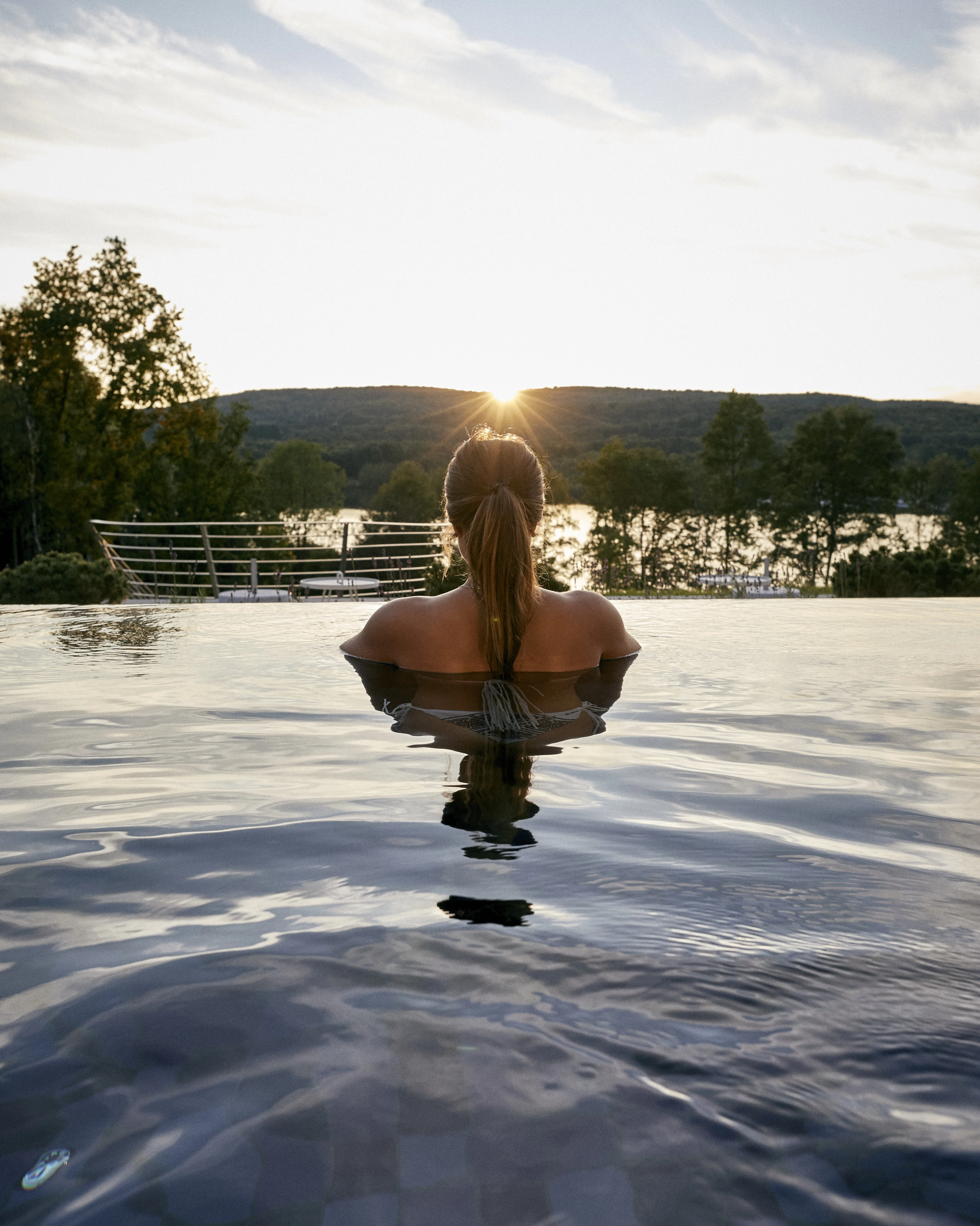 Frau entspannt in Infinity-Pool mit Sonnenuntergang und Landschaft im Hintergrund