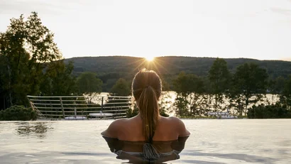 Frau entspannt in Infinity-Pool mit Sonnenuntergang und Landschaft im Hintergrund