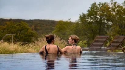 Zwei Frauen entspannen im Außenpool mit Blick auf Bäume und Hügel