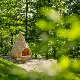 Gemütlicher Korbsessel mit Kissen im Grünen neben einem Steingehweg