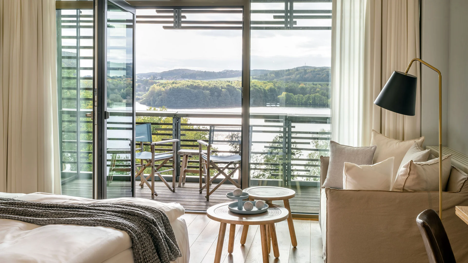 Hotelzimmer mit Balkon, Stühlen und Blick auf See und bewaldete Hügel