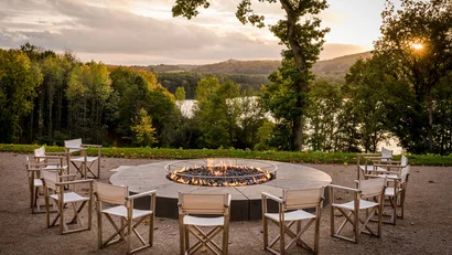 Stuhlkreis um ein Feuer im Freien mit Blick auf Wald und See bei Sonnenuntergang